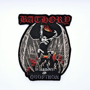 Könnte beinhalten: Gestickter Aufnäher mit dem Wort "BATHORY" in roten gotischen Buchstaben. Der Aufnäher zeigt eine geflügelte Figur mit Flammen und den Text "IN MEMORY OF QUORTHON" in Weiß.