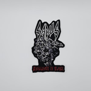Op de afbeelding: Zwarte geborduurde patch met het woord "SADUS" in witte, gestileerde letters. Daaronder een zwart-wit grafisch ontwerp. De tekst "SWALLOWED IN BLACK" is in het rood geborduurd aan de onderkant van de patch.