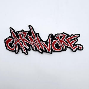 May include: A black embroidered patch with the word "CARNIVORE" in red, stylized lettering. The letters have a white outline and are on a black background. The patch is rectangular and has a jagged edge.