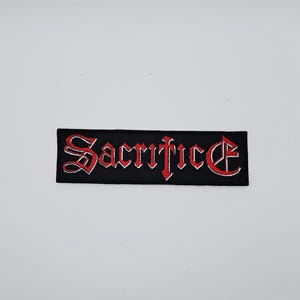 Puede incluir: Parche rectangular negro con la palabra "Sacrifice" bordada en letras góticas rojas. El parche tiene un borde rojo y está diseñado para ser cosido en ropa o accesorios.