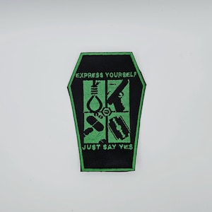 May include: A black and green patch in a coffin shape. The patch features the text "EXPRESS YOURSELF" at the top and "JUST SAY YES" at the bottom. Inside are four green graphic images: a flame, a gun, pills, and a razor blade.