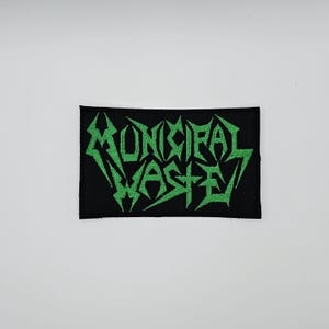 Może przedstawiać: Czarna prostokątna naszywka z napisem "MUNICIPAL WASTE" w jaskrawozielonych, stylizowanych, poszarpanych literach. Naszywka jest prawdopodobnie przeznaczona na kurtkę lub torbę, a tekst sugeruje zespół lub markę.
