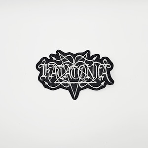 以下が含まれることがあります： ゴシック体で「TACONIA」と書かれた白黒の刺繍パッチ。パッチは五芒星と矢印のデザインで、ユニークな形をしています。