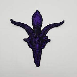 May include: A purple and black patch featuring a detailed illustration of a Baphomet head with a pentagram on its forehead. The patch has a black border and is set against a white background. The design is intricate and gothic in style.