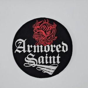 Può includere: Toppa rotonda nera con le parole "Armored Saint" in lettere gotiche bianche. Un'illustrazione rossa di un volto con elmo è sopra il testo. La toppa è probabilmente per una giacca o una borsa.