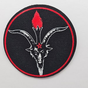 May include: A black and white embroidered patch with a red border. The patch features a goat's head with horns and a five-pointed star with a circle in the center. The goat's head is surrounded by a flame.