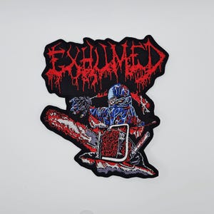 May include: Embroidered patch with the word "EXHUMED" in dripping red lettering. It depicts a figure in blue and red using a chainsaw. The patch has a black border. The design is on a white background.
