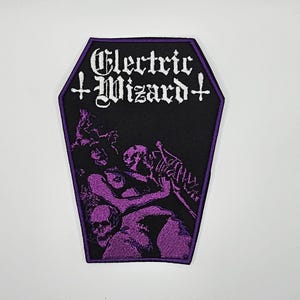 Puede incluir: Parche negro en forma de ataúd con las palabras "Electric Wizard" en letras góticas blancas. El parche presenta una ilustración púrpura de figuras y calaveras. El parche tiene un borde púrpura.