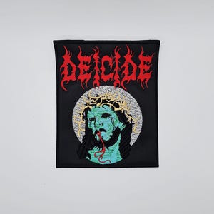 May include: Embroidered black patch featuring the word "DEICIDE" in red, stylized lettering. The patch depicts a figure with a crown of thorns, with teal skin and a red tongue. The figure is set against a white circular background.