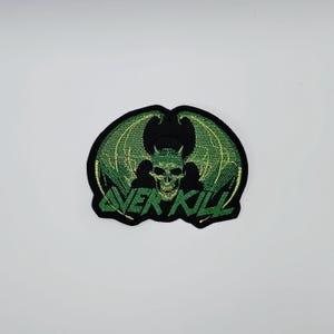 May include: Embroidered patch with a green skull and bat wings, and the text "OVER KILL" in green. The patch has a black border and is on a white background. Ideal for jackets or bags.