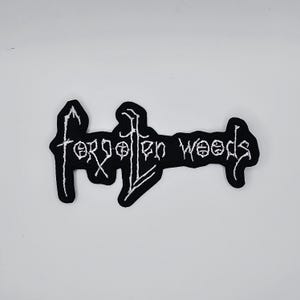 Può includere: Toppa ricamata in bianco e nero con la scritta "forgotten woods" in un carattere gotico. La toppa ha uno sfondo nero e cuciture bianche. La toppa ha la forma di una forma stilizzata.