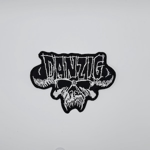 Może przedstawiać: Czarna i biała naszywka z haftem z napisem "DANZIG" w gotyckiej czcionce nad czaszką z rogami. Naszywka ma nieregularny kształt i jest przeznaczona do odzieży lub akcesoriów.