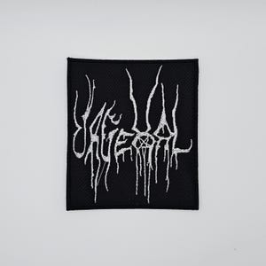 Puede incluir: Parche cuadrado negro con texto blanco bordado. El texto, de estilo gótico, parece ser el nombre de una banda o un logotipo, con un pentagrama. El diseño tiene un efecto de goteo.