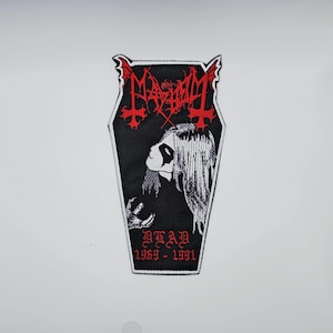 Puede incluir: Parche negro en forma de ataúd con detalles bordados en rojo y blanco. Presenta el nombre de la banda "Mayhem" en letras góticas rojas, un retrato de una persona con pintura facial blanca y el texto "DEAD 1969-1991".