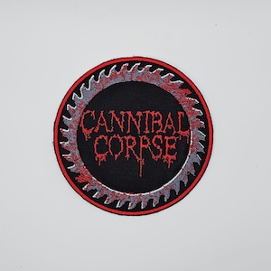 May include: A round, black patch with the text "CANNIBAL CORPSE" in red, dripping letters. The patch has a red border and a gray saw blade design around the text.
