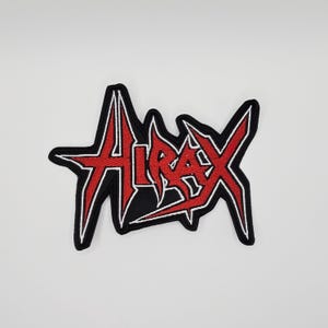 Può includere: Toppa ricamata con la scritta "Anthrax" in rosso, lettering stilizzato con contorno bianco, su sfondo nero. La toppa ha una forma frastagliata a stella.