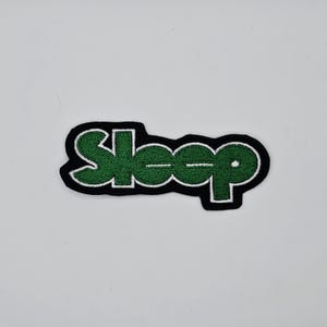 Puede incluir: Parche bordado con la palabra "Sleep" en verde, contorneada en blanco y bordeada en negro. El parche rectangular está diseñado para planchar o coser sobre tela.
