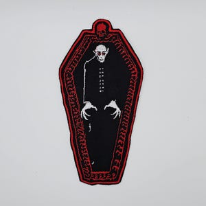 Peut inclure: Écusson brodé représentant un vampire dans un cercueil. Le patch a un fond noir avec un contour de cercueil rouge et un crâne en haut. Le vampire est blanc avec des yeux rouges et porte un costume noir.