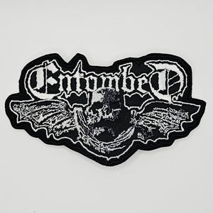 May include: A black and white embroidered patch featuring the text Entombed in a stylized font above a central skull graphic with wings extending to the sides. The design is outlined in black thread with a detailed white stitched texture.