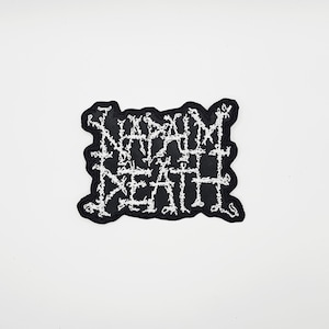 Puede incluir: Un parche bordado en blanco y negro con el texto "NAPALM DEATH" en una fuente gótica estilizada. El parche tiene un borde negro y está diseñado para ser cosido o planchado en ropa o accesorios.
