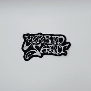 May include: A black and white embroidered patch with the text "MORBID SAINT" in a stylized font. The patch has a black border and the text is in white. The patch is on a white background.