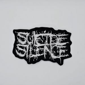 Könnte beinhalten: Schwarzer, gestickter Aufnäher mit dem Text "SUICIDE SILENCE" in weißer, stilisierter, gotischer Schrift. Der Aufnäher hat einen gezackten, unregelmäßigen Rand. Der Aufnäher ist wahrscheinlich für eine Jacke oder Tasche.