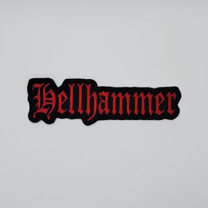 May include: A black embroidered patch with the word "Hellhammer" in red gothic lettering. The patch has a black border and is likely designed to be sewn or ironed onto clothing or accessories.