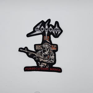 Peut inclure: Écusson brodé noir et blanc avec le mot "Sodom" au-dessus d'une croix. En dessous, une illustration détaillée d'une figure tenant un fusil, avec le texte "PERSECUTION MANIA" en rouge.