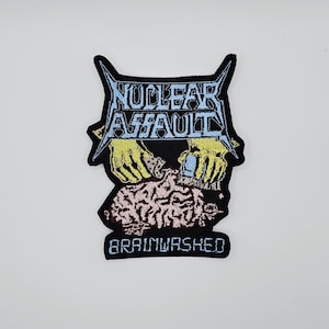Puede incluir: Parche bordado con las palabras "NUCLEAR ASSAULT" en una fuente estilizada, sobre un gráfico de manos trabajando en un cerebro. La palabra "BRAINWASHED" está en la parte inferior. El parche es negro con detalles amarillos, azules y rosas.