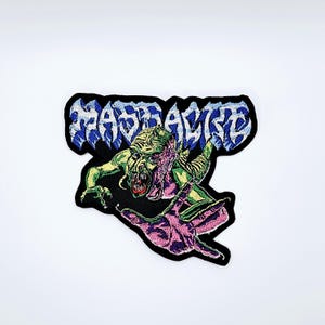 May include: Embroidered patch featuring a green and purple monster with the word "MASSACRE" in blue and white text. The monster has a menacing expression and is reaching out with a pink hand. The patch has a black border.