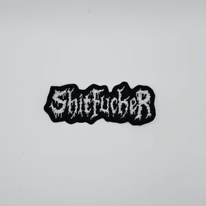 May include: A black and white embroidered patch featuring the word "Shitfucker" in a gothic font. The patch has a black border and is set against a white background. The text is in a stylized, distressed font.