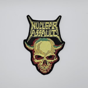 May include: Embroidered patch with a skull and the text "NUCLEAR ASSAULT" in yellow. The skull is yellow and red, with green details. The patch has a black border. Suitable for application on clothing or accessories.