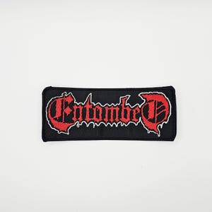 May include: Black rectangular patch with the word "Entombed" in red gothic lettering, outlined in white. The patch is likely for a band or music-related merchandise.
