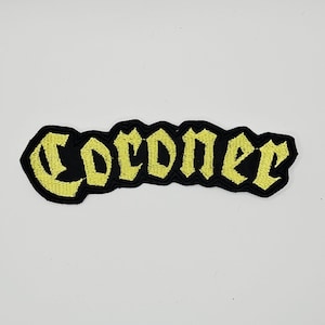 May include: An embroidered patch featuring the word Coroner in a yellow gothic-style font with a thick black border. The lettering is stitched with a textured thread finish, creating a bold contrast against the black fabric background of the patch.