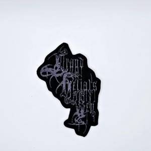 May include: Black embroidered patch with the text "Thy Rituals Weep" in a gothic font. The patch has a unique, irregular shape and features gray thread detailing. Ideal for jackets, bags, or other fabric items.