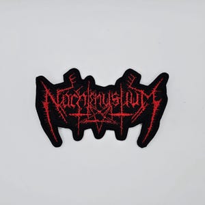 May include: Black embroidered patch with the word "Nacrentuslume" in red, gothic lettering. The patch features a pentagram and other symbols. The patch is likely for a jacket or bag.
