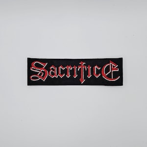 May include: Black rectangular patch featuring the word "Sacrifice" in red gothic lettering with a white outline. The patch is designed for application on clothing or accessories.
