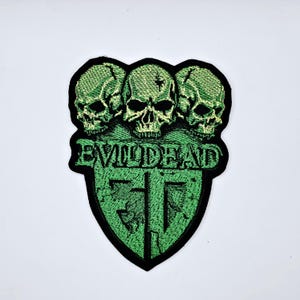 Puede incluir: Parche bordado con tres calaveras verdes sobre el texto "EVIL DEAD" y un diseño de escudo verde. El parche tiene un borde negro, ideal para ropa o accesorios.