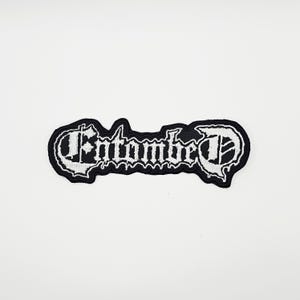 Puede incluir: Parche bordado en blanco y negro con la palabra "Entombed" en una fuente gótica. El parche tiene un borde negro con un borde ondulado y está sobre un fondo blanco.