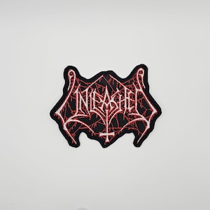 May include: Embroidered patch with the word "UNLEASHED" in a gothic font, outlined in white, over a black background. The design includes a red web-like pattern and a cross symbol at the bottom. The patch has a unique, irregular shape.