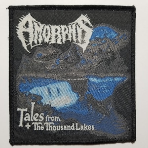May include: A black woven patch with the band name "AMORPHIS" in white lettering. The patch features a blue and gray landscape with the text "Tales from The Thousand Lakes" in white lettering.