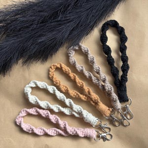 May include: A selection of macrame wrist lanyards in shades of pink, white, beige, and black. Each lanyard has a twisted rope pattern and a silver-tone clasp. A dark, feathery plant is visible in the background.