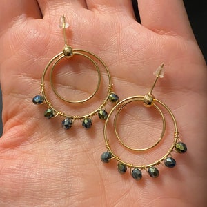 Puede incluir: Pendientes de aro dorados con dos círculos y cuentas negras que cuelgan de la parte inferior.