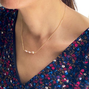 May include: A delicate gold chain necklace with three small pearl accents. The necklace is worn by a person with a floral patterned shirt.