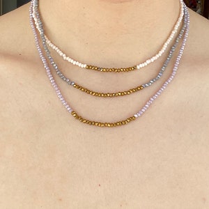May include: A layered necklace with four strands of beads. The top strand is white, the second is gray, the third is light purple, and the bottom strand is gold.