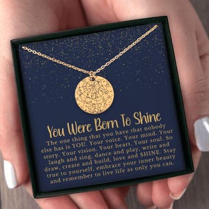 May include: A gold-toned necklace with a circular pendant featuring a star map design. The necklace is presented in a black box with a dark blue card printed with gold stars and the text: You Were Born To Shine, followed by an inspirational message.