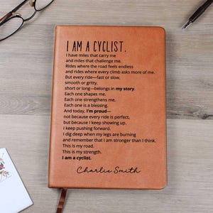 May include: A brown leather journal with the text "I AM A CYCLIST" and a poem about cycling. The journal has a ribbon bookmark and is on a wooden surface with glasses, a pen, and a card.