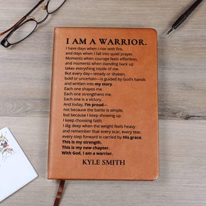 May include: A brown leather journal with the text "I AM A WARRIOR" and a poem printed on the cover. The journal has a ribbon bookmark and is surrounded by a pen, glasses, and a card.