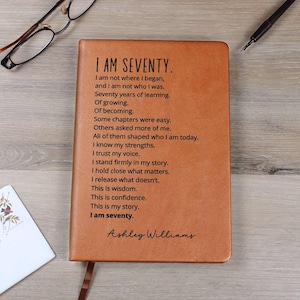 May include: A tan leather journal with the text "I AM SEVENTY" and a poem printed on the cover. The journal has a ribbon bookmark and the name "Ashley Williams" at the bottom. Glasses and a pen are on the wooden surface.