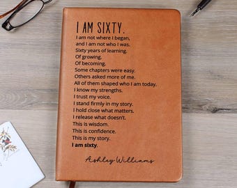 May include: A brown leather journal with the text "I AM SIXTY" and a poem about life experiences. The journal has a ribbon bookmark and the name "Ashley Williams" at the bottom. Glasses and a pen are visible.